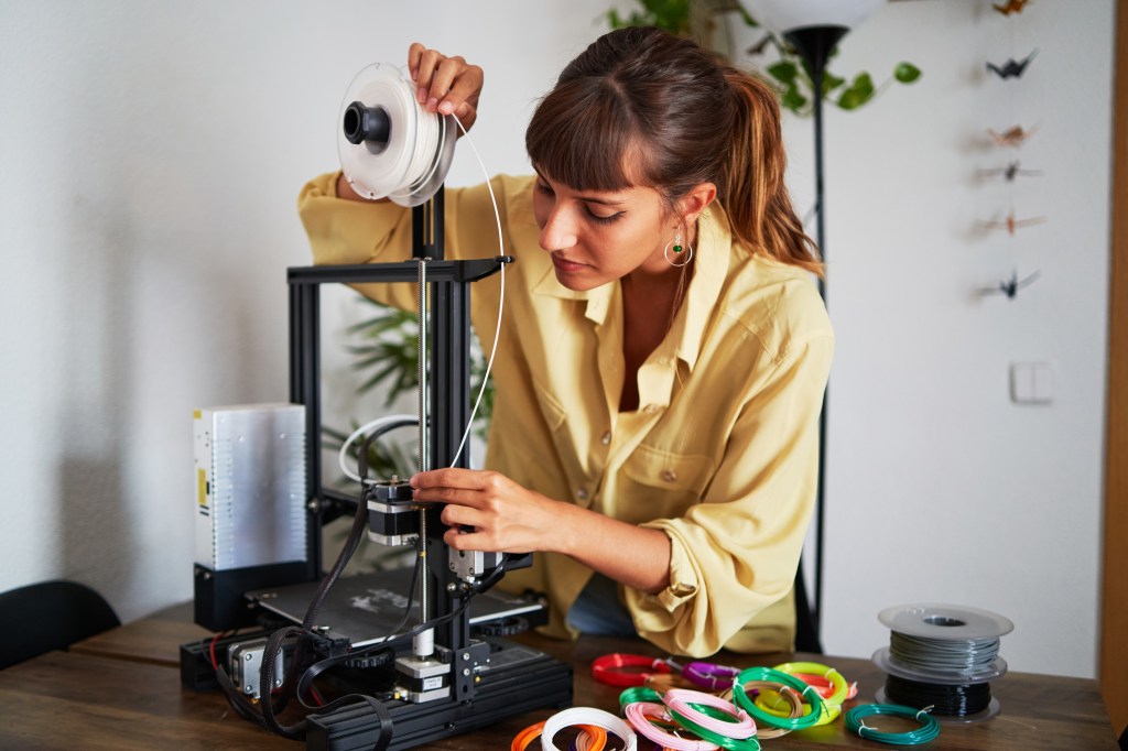 Portrait of a confident young woman standing while working at home with her 3d printer while preparing the filaments inside printer.
